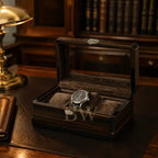 A dark wood watch storage box with a clear glass lid, velvet interior, and silver latch, shown open on a wooden desk with a watch inside. A brass desk lamp and bookshelves are visible in the background.