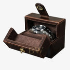 A dark brown leather watch box with a hinged lid and pull tab, shown open to reveal a silver-toned metal watch inside on a light gray background.