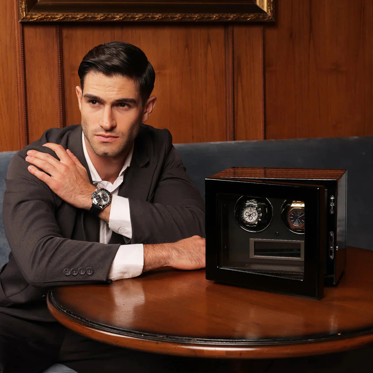 British man in a suit with a watch on his wrist next to a watch winder on a wooden table.