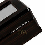 Close-up of a dark wood watch box with a glossy finish, showing a glass window and silver metal hinge details on a white background.