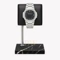 A silver-toned stainless steel Patek Philippe Nautilus with a black dial and date window, displayed on a black cylindrical stand mounted over a black marble base, against a light gray background.