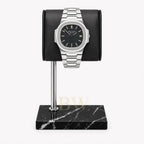 A silver-toned stainless steel Patek Philippe Nautilus with a black dial and date window, displayed on a black cylindrical stand mounted over a black marble base, against a light gray background.