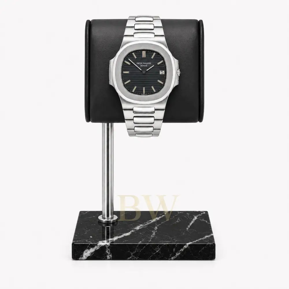 A silver-toned stainless steel Patek Philippe Nautilus with a black dial and date window, displayed on a black cylindrical stand mounted over a black marble base, against a light gray background.