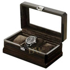 A luxury watch box with a dark wood grain exterior, a clear glass lid, and a plush dark brown velvet interior. A silver-tone chronograph watch with a black strap is displayed inside one of the three watch slots.