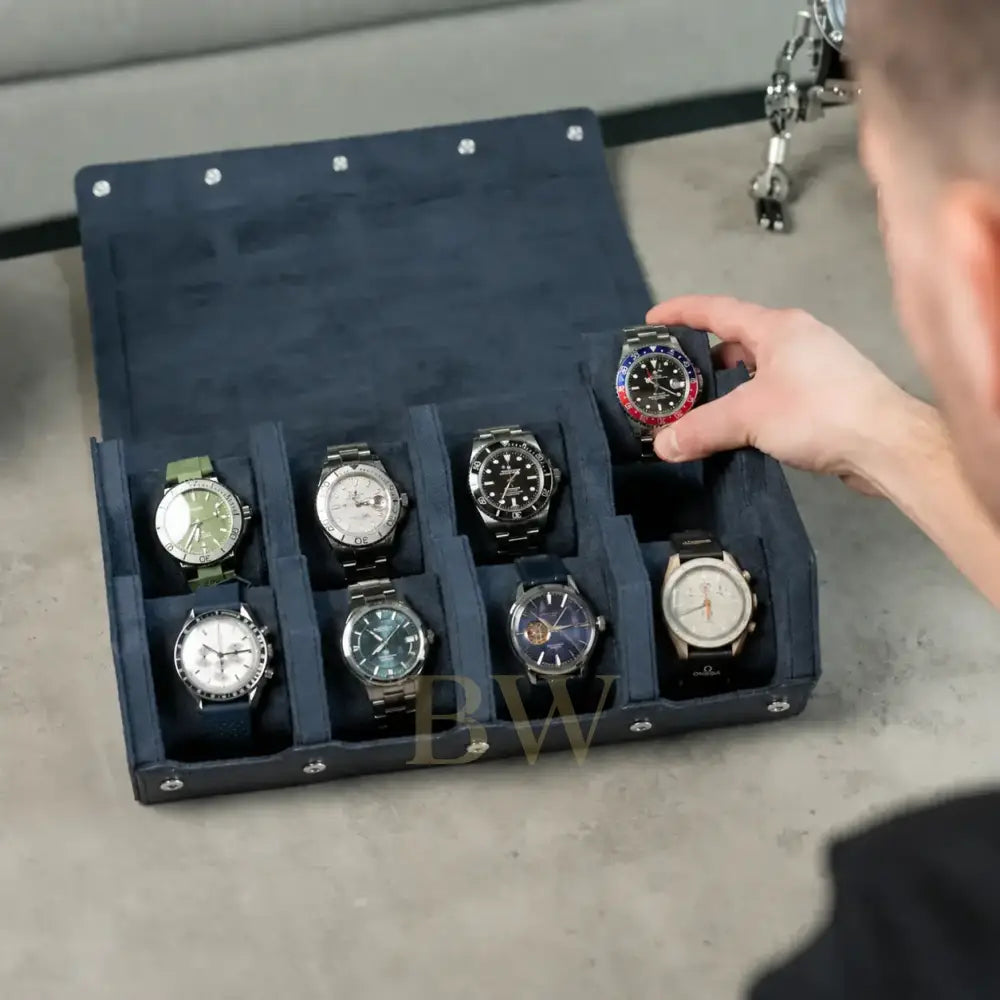 A navy blue watch storage roll with eight compartments, shown open on a concrete floor. Multiple wristwatches are arranged inside, and a person's hand is reaching in to adjust or select one watch.