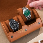 A tan leather watch roll with plush brown suede-like compartments holding two wristwatches, shown with a hand placing or adjusting the right watch. The product being sold appears to be the watch roll organizer rather than the watches themselves, since the rolls and exterior handle are prominently featured and the watches function as demonstration props.