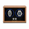 A wooden dual watch winder with a medium brown wood-grain finish, black textured interior, and two circular watch slots illuminated by blue LED lights. Two stainless Rolex and Audemars Piguet are displayed inside.