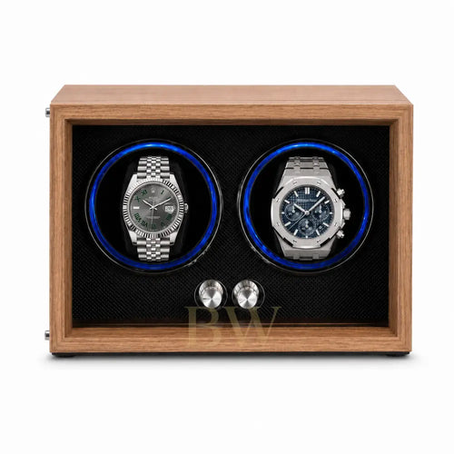 A wooden dual watch winder with a medium brown wood-grain finish, black textured interior, and two circular watch slots illuminated by blue LED lights. Two stainless Rolex and Audemars Piguet are displayed inside.