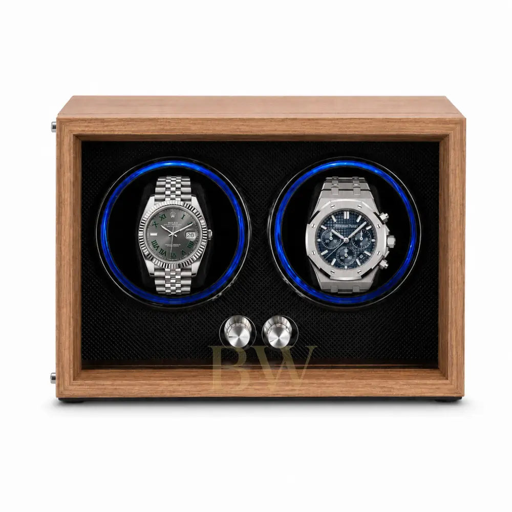 A wooden dual watch winder with a medium brown wood-grain finish, black textured interior, and two circular watch slots illuminated by blue LED lights. Two stainless Rolex and Audemars Piguet are displayed inside.